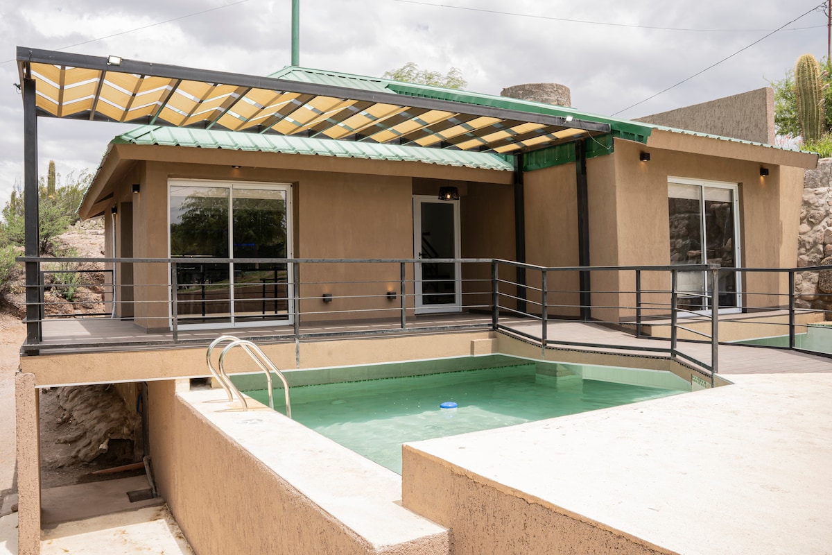 A modern exterior features a covered patio area leading to a private pool. Large glass doors provide natural light and easy access to the pool area. The structure is surrounded by minimal landscaping, emphasizing the tranquility of the setting.