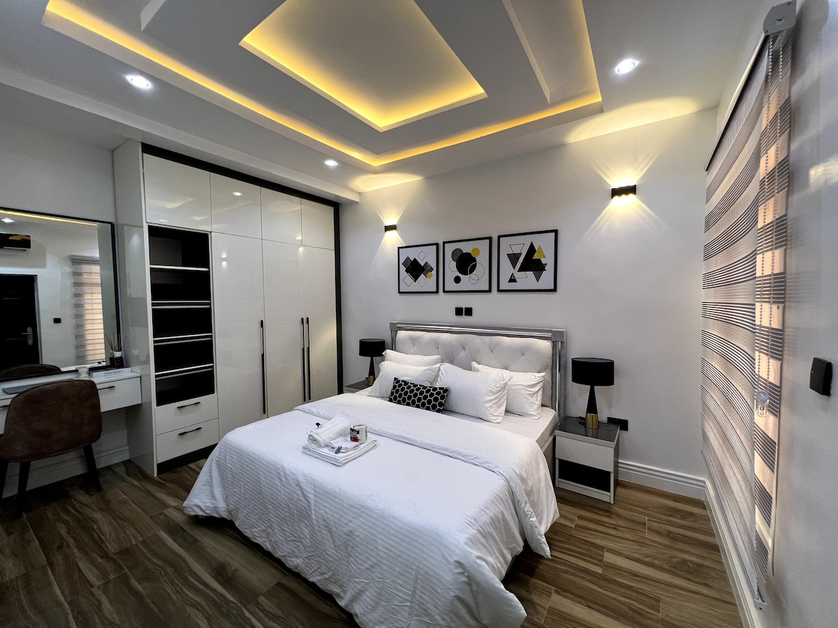 A modern bedroom features a spacious bed with smooth white linens and decorative pillows. Adjacent, a sleek wardrobe offers ample storage. Artwork in geometric designs adorns the walls, while soft, ambient lighting highlights the tray ceiling. A window with elegant blinds allows natural light to enter.