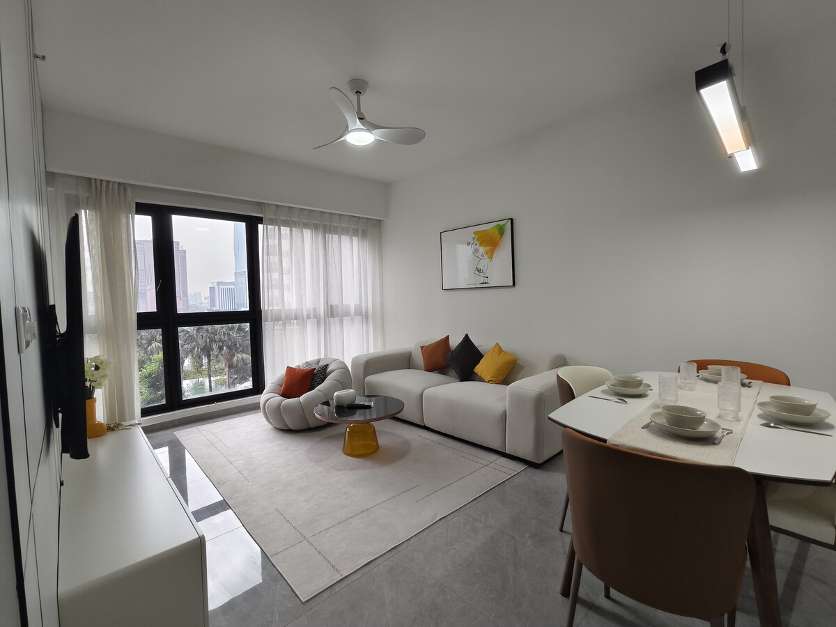 A modern living space showcases a minimalist design with a comfortable sofa and accent pillows. A stylish dining table is set for four, with tableware neatly arranged. Large windows invite natural light, complementing the light-colored walls and soft furnishings.