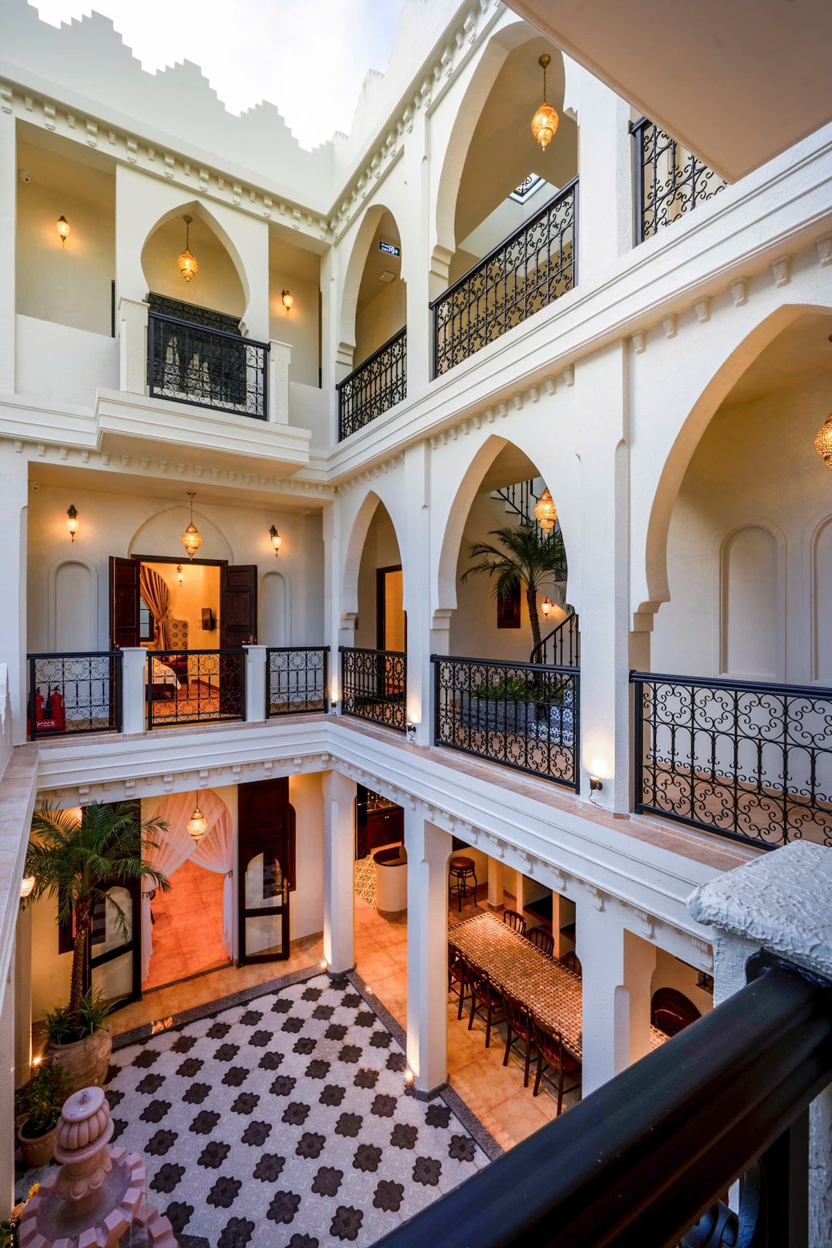 An open courtyard features elegant arches and intricate metalwork, creating a sense of spaciousness. Soft lighting highlights the architectural details, while a patterned tile floor provides visual interest. Potted palms add a touch of greenery, enhancing the tranquil ambiance of the space.