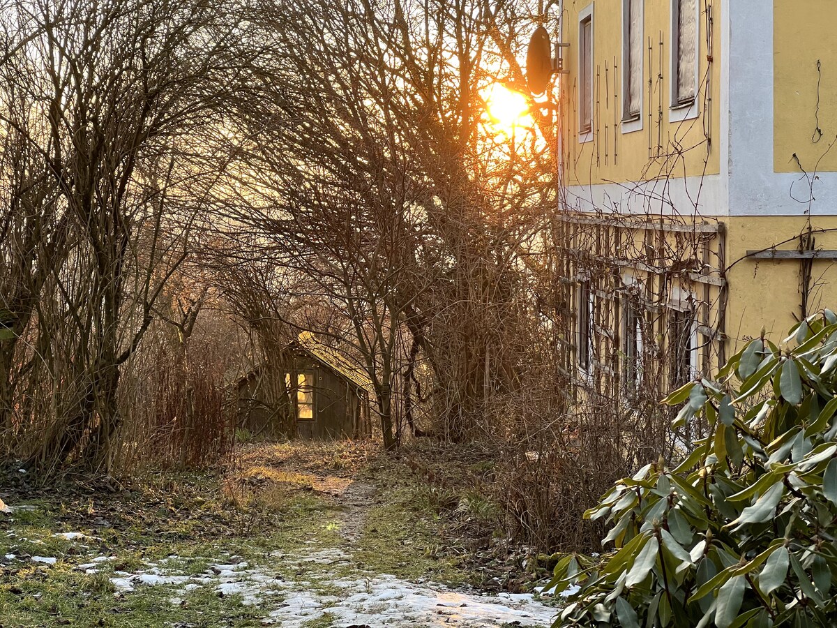 A sunlit path meanders beside the yellow exterior of the building, framed by bare trees and shrubbery. The warm glow of the sunset casts soft light over the scene, enhancing the serene atmosphere of the surrounding landscape.