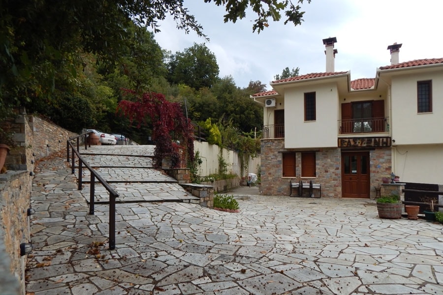 A stone-paved pathway leads to a charming two-story building surrounded by greenery. The exterior features a blend of natural stone and traditional architecture. A small set of stairs ascends alongside, and seating areas with potted plants are visible, inviting outdoor relaxation.