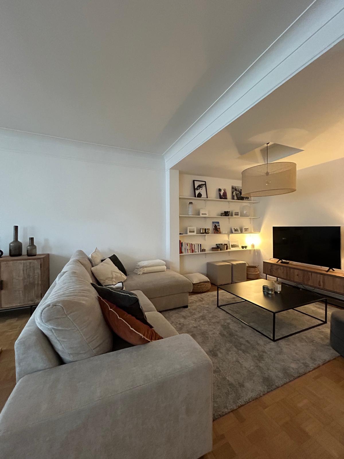 A spacious living area is presented with a gray rug and light-colored sofas. A coffee table sits in front of a media unit, which features a large television. Decorative shelves are mounted on the wall, exhibiting books and personal items, while soft lighting creates a warm ambiance.