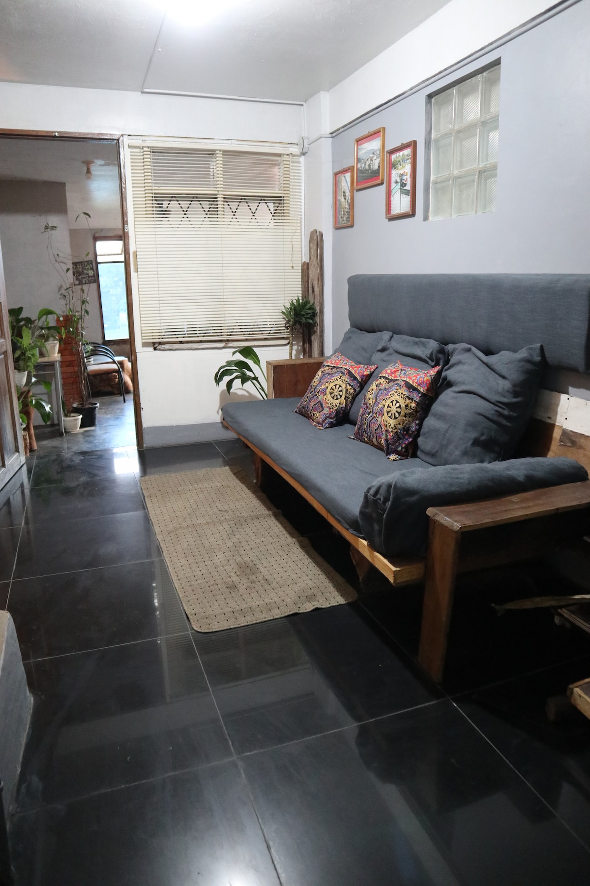 A narrow hallway is featured with polished black tile flooring and ample natural light from a nearby window. A wooden sofa with decorative cushions is positioned against the wall, complemented by a woven rug beneath. Potted plants add a touch of greenery to the space.