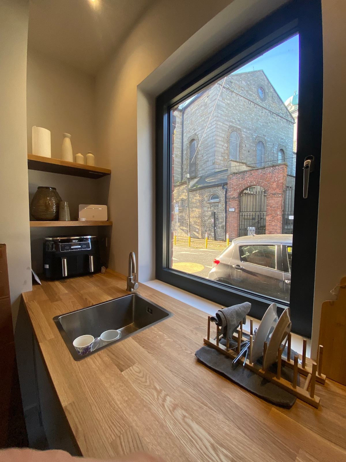 A modern kitchen area features a wooden countertop and a stainless steel sink, complemented by a window offering views of the street outside. Nearby, a coffee maker and several white dishware items are arranged. A dish rack holds various plates and utensils.