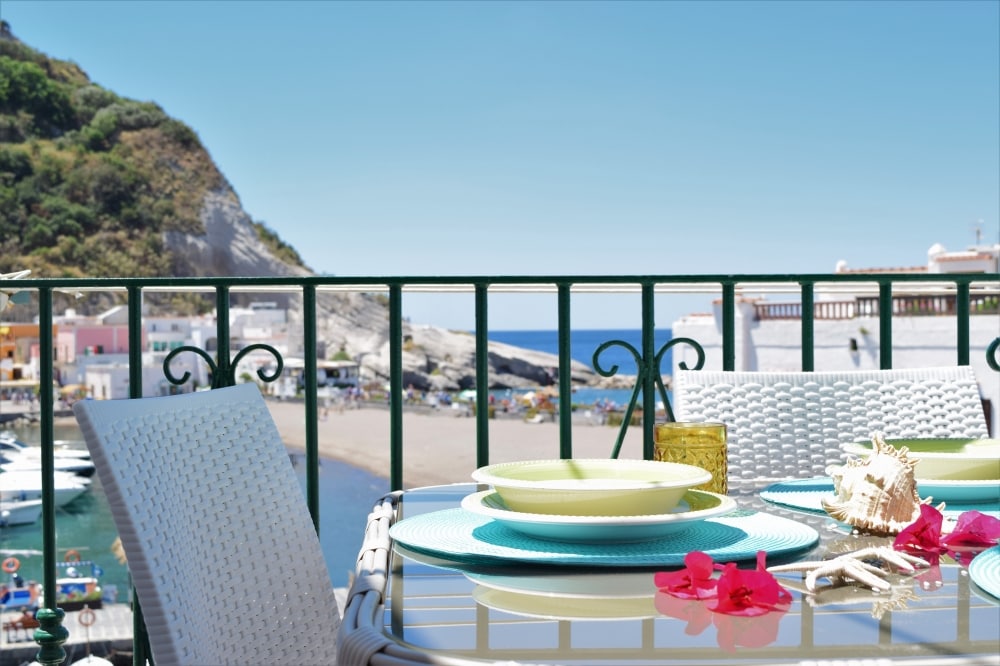 A dining table is set with colorful plates and a glass, accompanied by tropical flowers and a seashell. The beautiful coastal view showcases the beach and vibrant buildings nestled against a rocky hill, with the sea visible in the background.