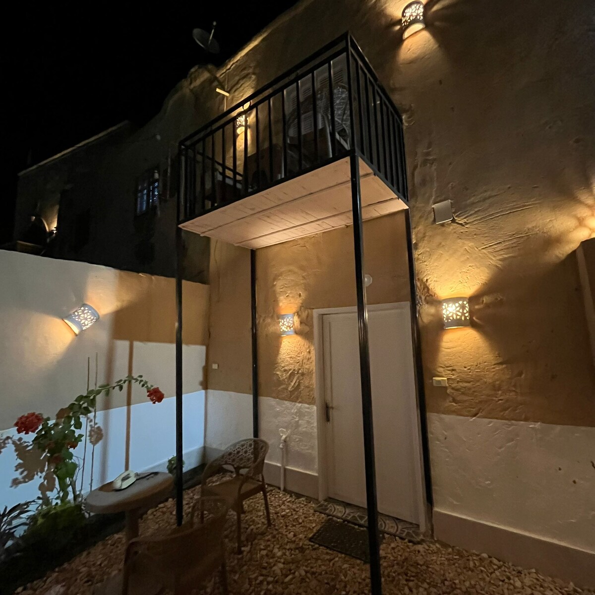 A charming courtyard is illuminated by warm, soft lighting. A small balcony is visible above the entrance, supported by sleek black railings. Two outdoor chairs rest on a pebbled surface, accompanied by potted plants, creating a serene space for relaxation.