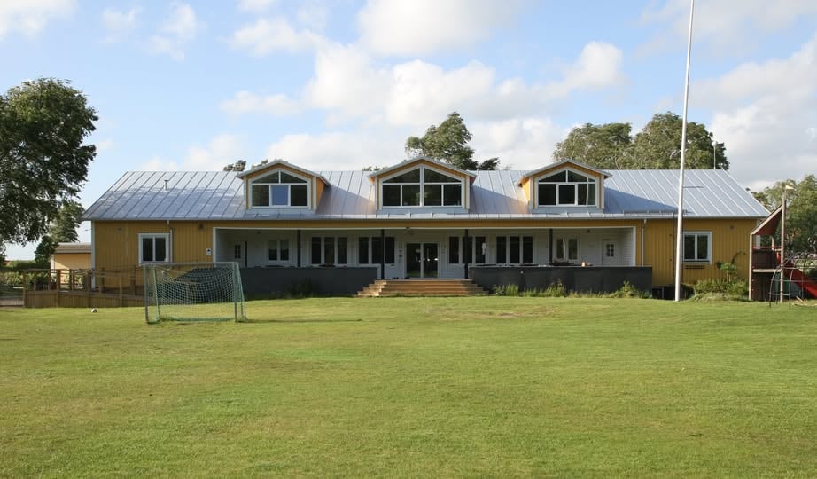 A spacious building is set against a backdrop of greenery, featuring a light wooden exterior and a metal roof. Large windows allow natural light into the interior. The surrounding area includes an open lawn and a soccer goal, creating an inviting outdoor space.