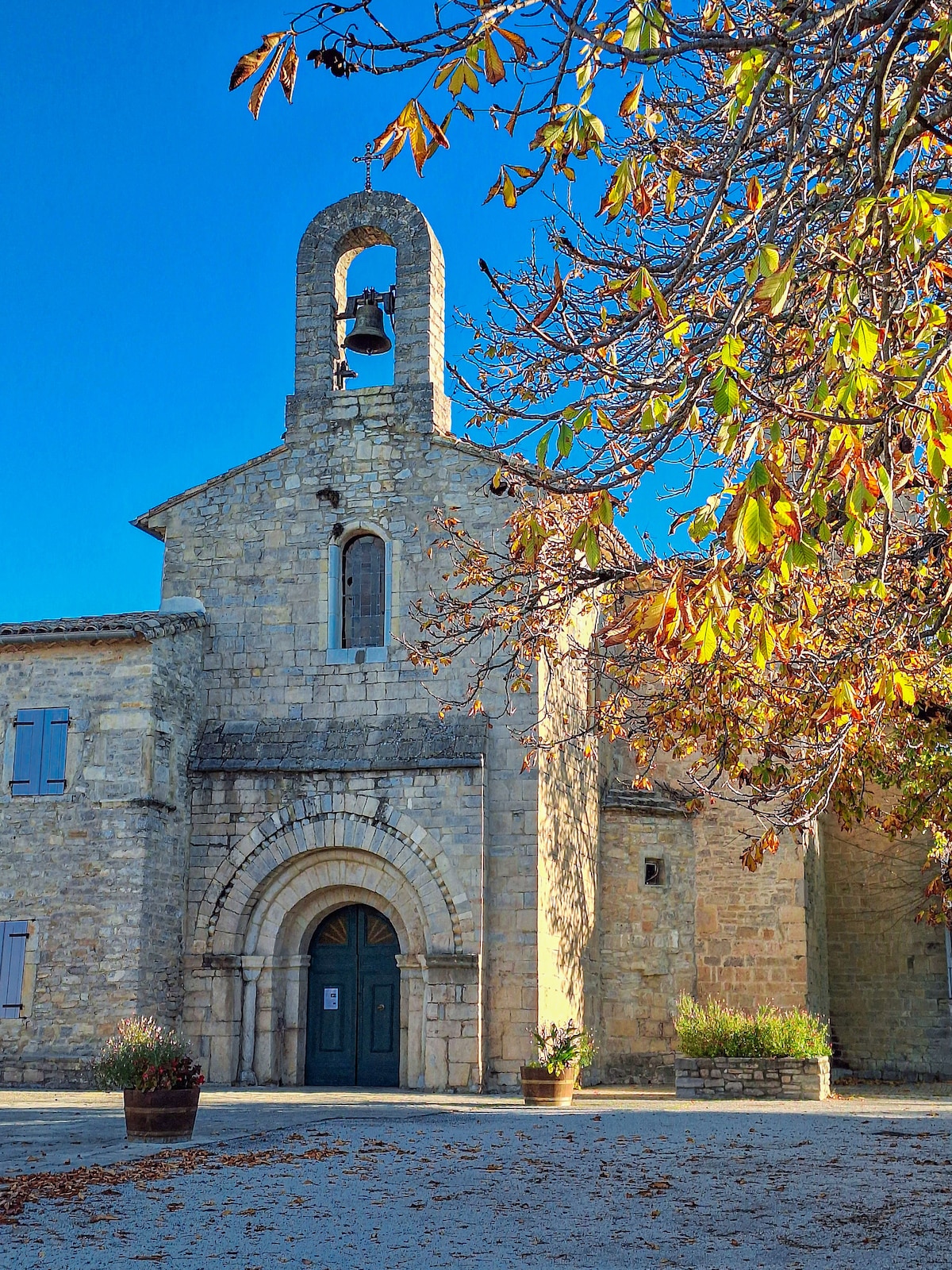 A historic stone building is featured, showcasing a charming bell tower and a large arched doorway. Surrounding foliage displays autumn colors, adding warmth to the scene. Clear blue skies provide a contrasting backdrop, enhancing the structure's details.