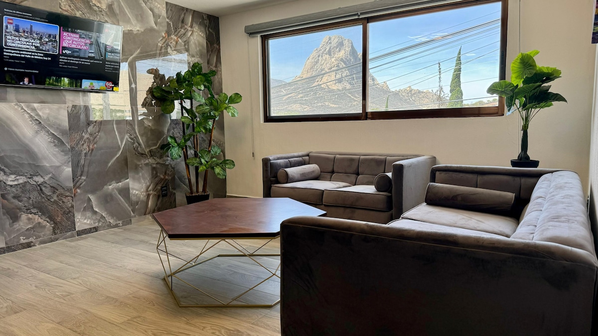 A contemporary living area features two comfortable sofas arranged around a low coffee table, with a large window providing a view of the Peña. Lush green plants are placed beside the seating, while a wall-mounted television adds entertainment options.