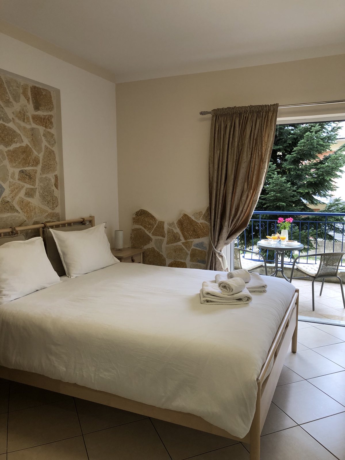 A comfortable sleeping area features a spacious bed with crisp white linens. Natural light floods in from a balcony door, which leads to an outdoor seating area with a small table and chairs. Soft, neutral tones are complemented by a textured stone wall.