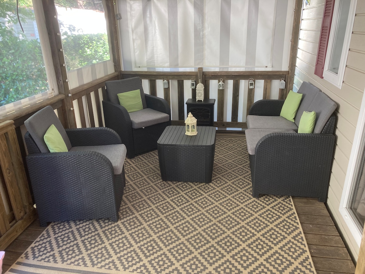 A cozy seating area is presented on a covered porch, featuring two armchairs and a loveseat with gray cushions and green accents. A small coffee table sits in the center. The space is enclosed by sheer curtains and showcases a patterned rug on the wooden floor.