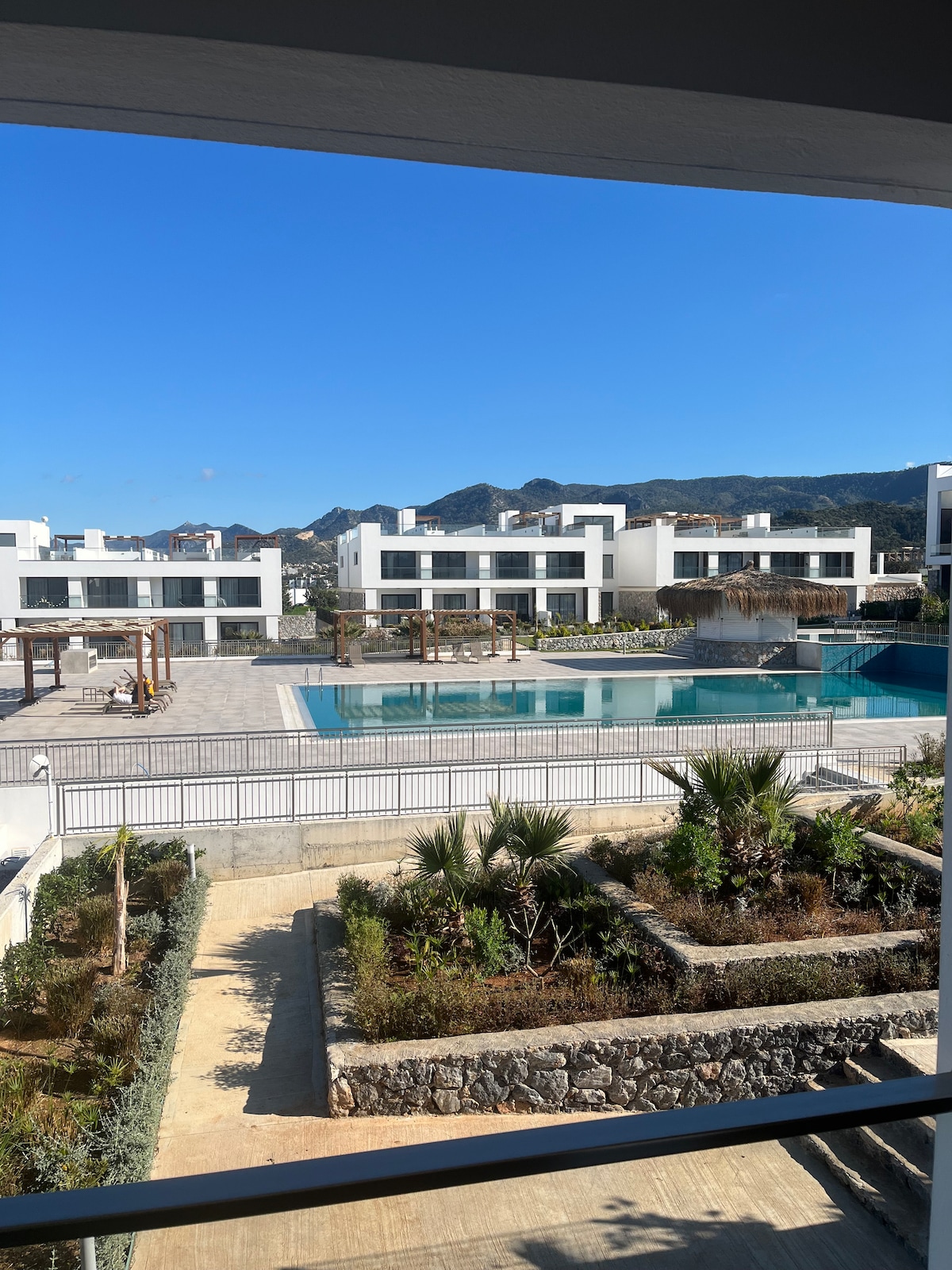 A view from the balcony reveals an outdoor area featuring a large swimming pool surrounded by a spacious deck. Green plantings and landscaped areas are visible in the foreground, with modern white buildings positioned against a backdrop of mountains under a clear blue sky.
