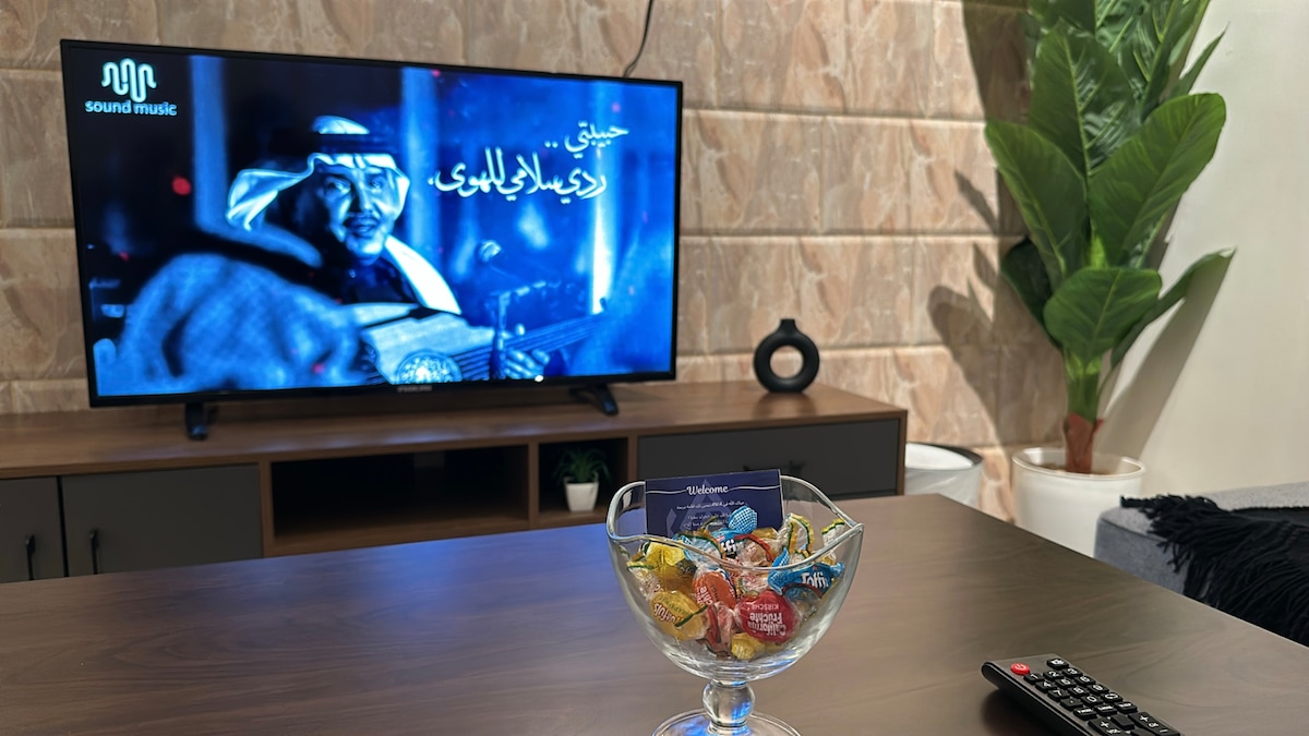 A modern living area is depicted, featuring a sleek television displaying entertainment options. A decorative glass bowl filled with colorful chocolates is positioned on a dark coffee table. A green potted plant adds a touch of nature, complementing the neutral tones of the space.