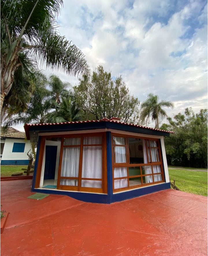 The exterior of a charming cottage displays large windows framed in wood, allowing natural light to fill the interior. A vibrant red patio floor encircles the building, accompanied by lush greenery and palm trees, creating a serene setting for relaxation.