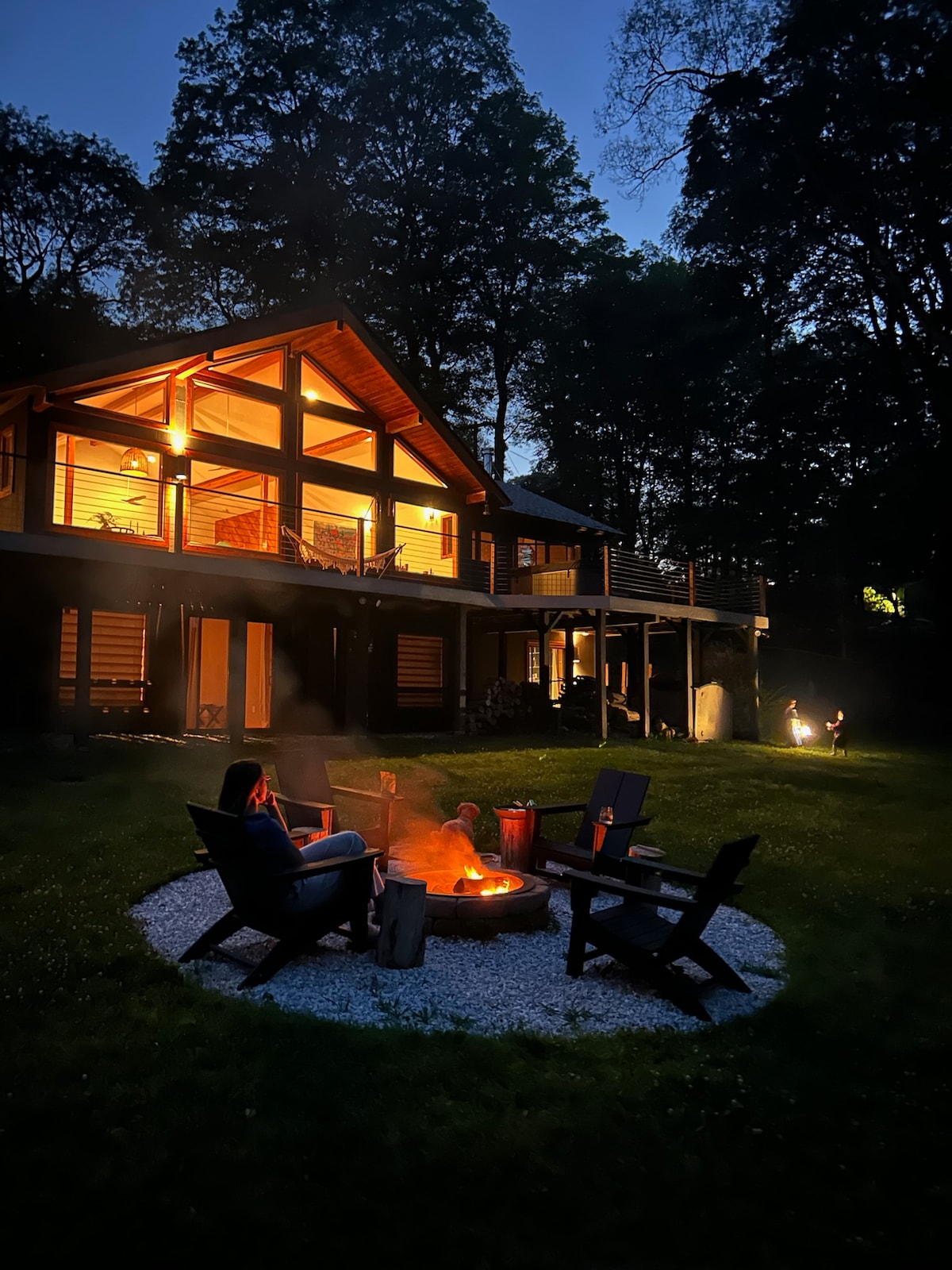 A spacious outdoor area features a fire pit surrounded by four chairs, with a grassy lawn leading to a well-lit house. The structure is partially illuminated from within, and trees frame the scene, creating a serene evening ambiance.