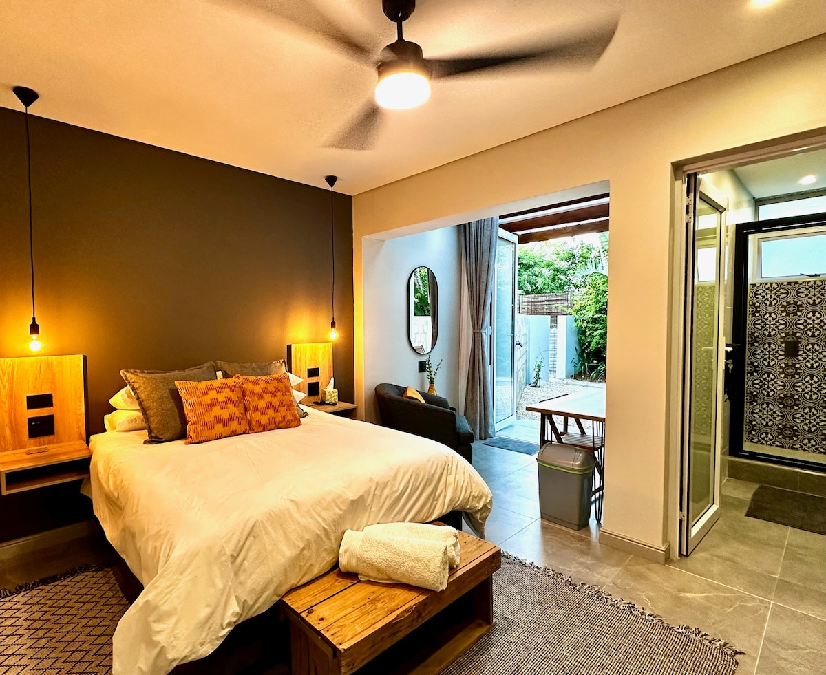 A spacious bedroom features a large bed with a variety of pillows. Natural light is allowed in through a sliding door, leading to an outdoor area. A fan is mounted to the ceiling, and the adjacent bathroom displays decorative tile work.