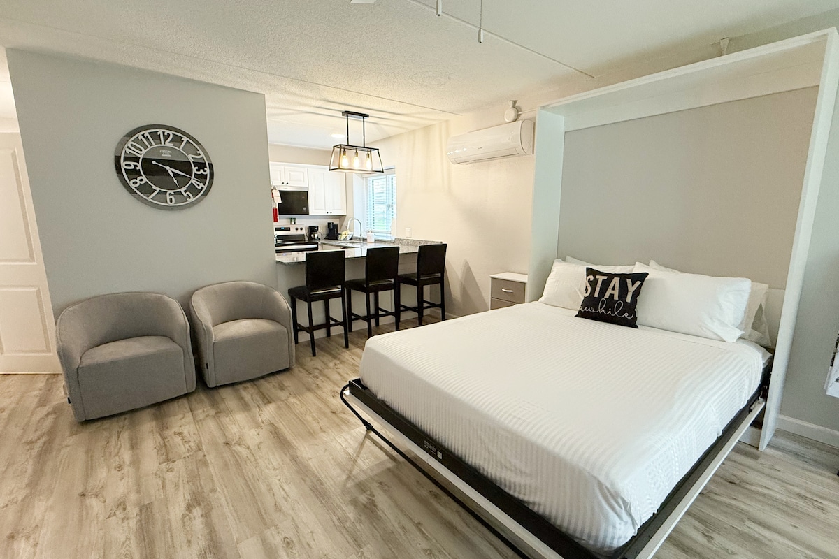 A open living space is seen, featuring a queen-size Murphy bed that is neatly folded away. Two grey armchairs are positioned across from a wall-mounted clock. The kitchen area, with modern bar seating, is visible in the background, illuminated by pendant lights.