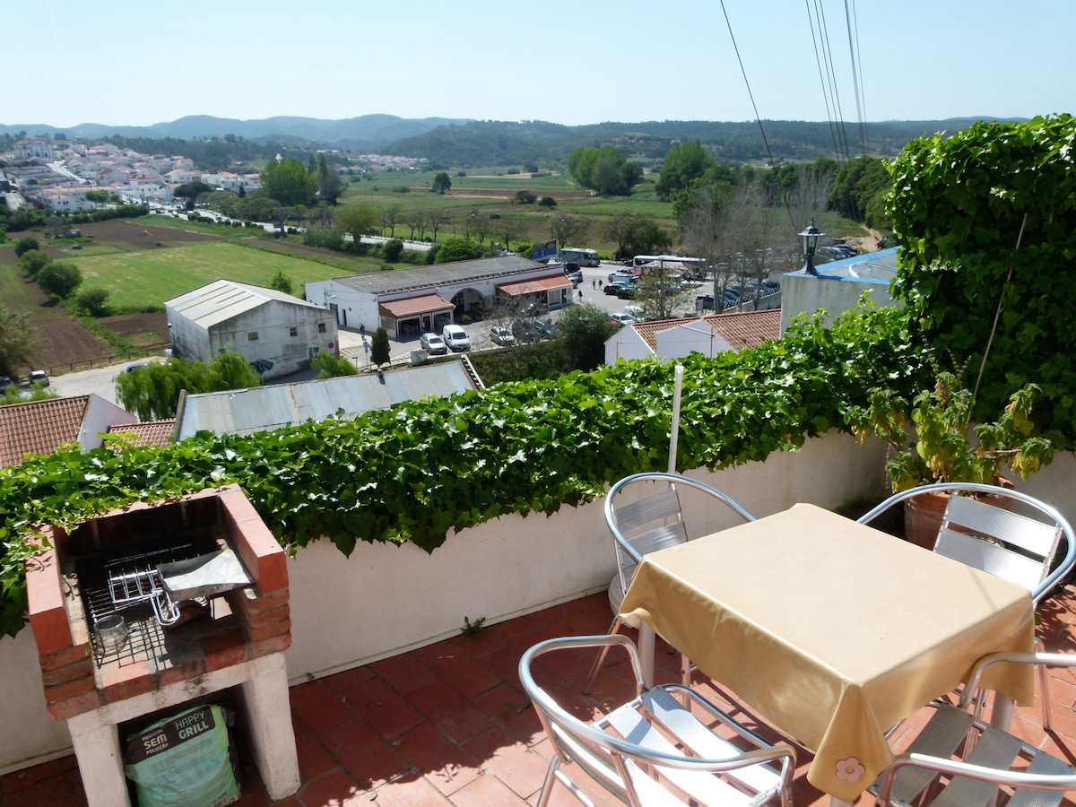 A terrace is shown, featuring a small table with four chairs, surrounded by lush green vines. A grill is present, and views of rolling hills and fields are visible in the background, creating an inviting outdoor space.