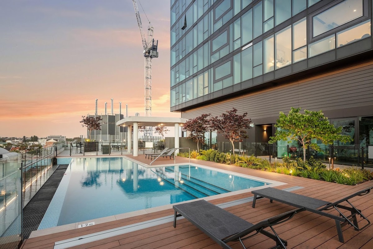 A rooftop pool area is featured, bordered by modern architecture. Comfortable lounge chairs are positioned on a wooden deck. Lush greenery and trees enhance the space, with a construction crane visible in the background. The scene is illuminated by the warm hues of sunset.