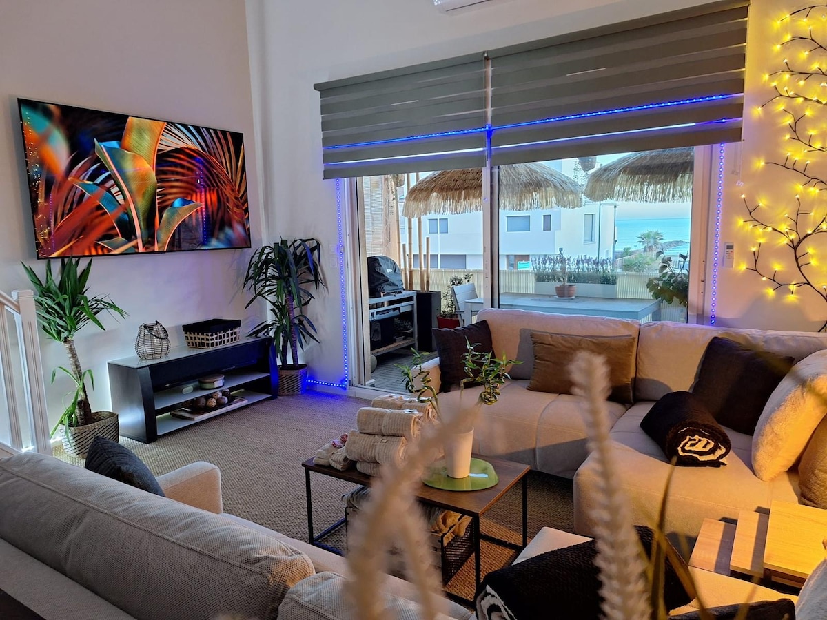 A modern living space features a large sectional sofa adorned with neutral-toned cushions. A vibrant television display showcases botanical imagery. Natural light enters through spacious glass doors that lead to an outdoor terrace with a beach view. Decorative plants enhance the inviting ambiance.