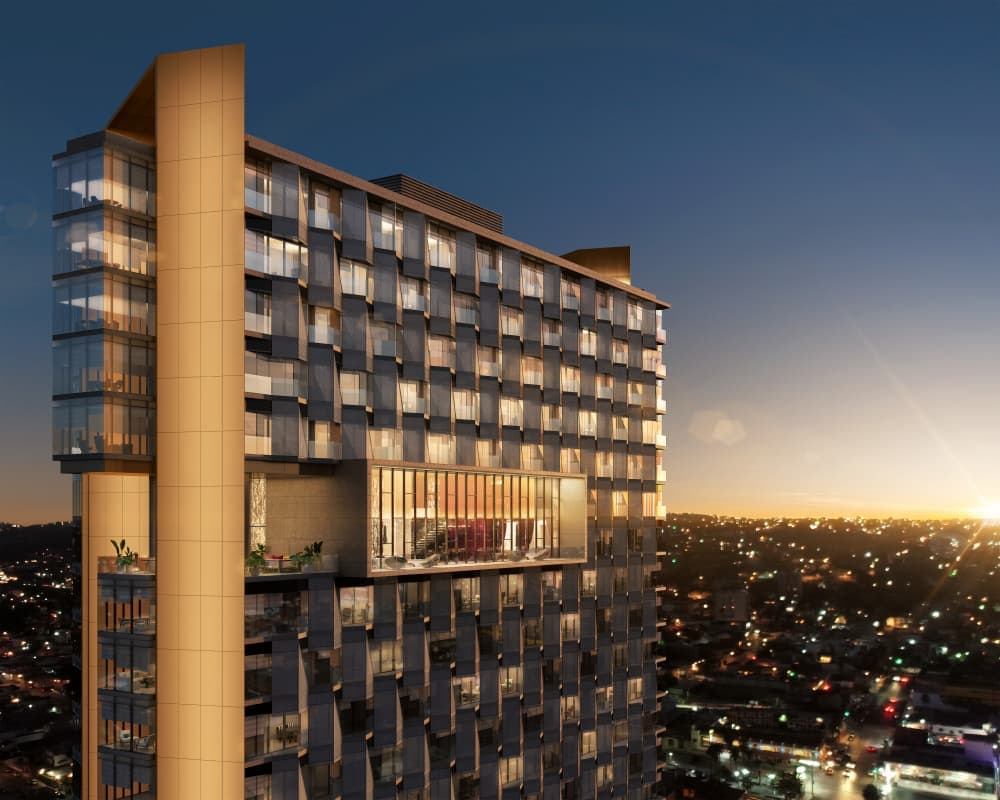 A modern high-rise building showcases sleek glass and metal façades, reflecting the evening light. A prominent balcony extends from the structure, revealing a spacious area for gatherings. The urban landscape below features twinkling city lights against a backdrop of dusk.
