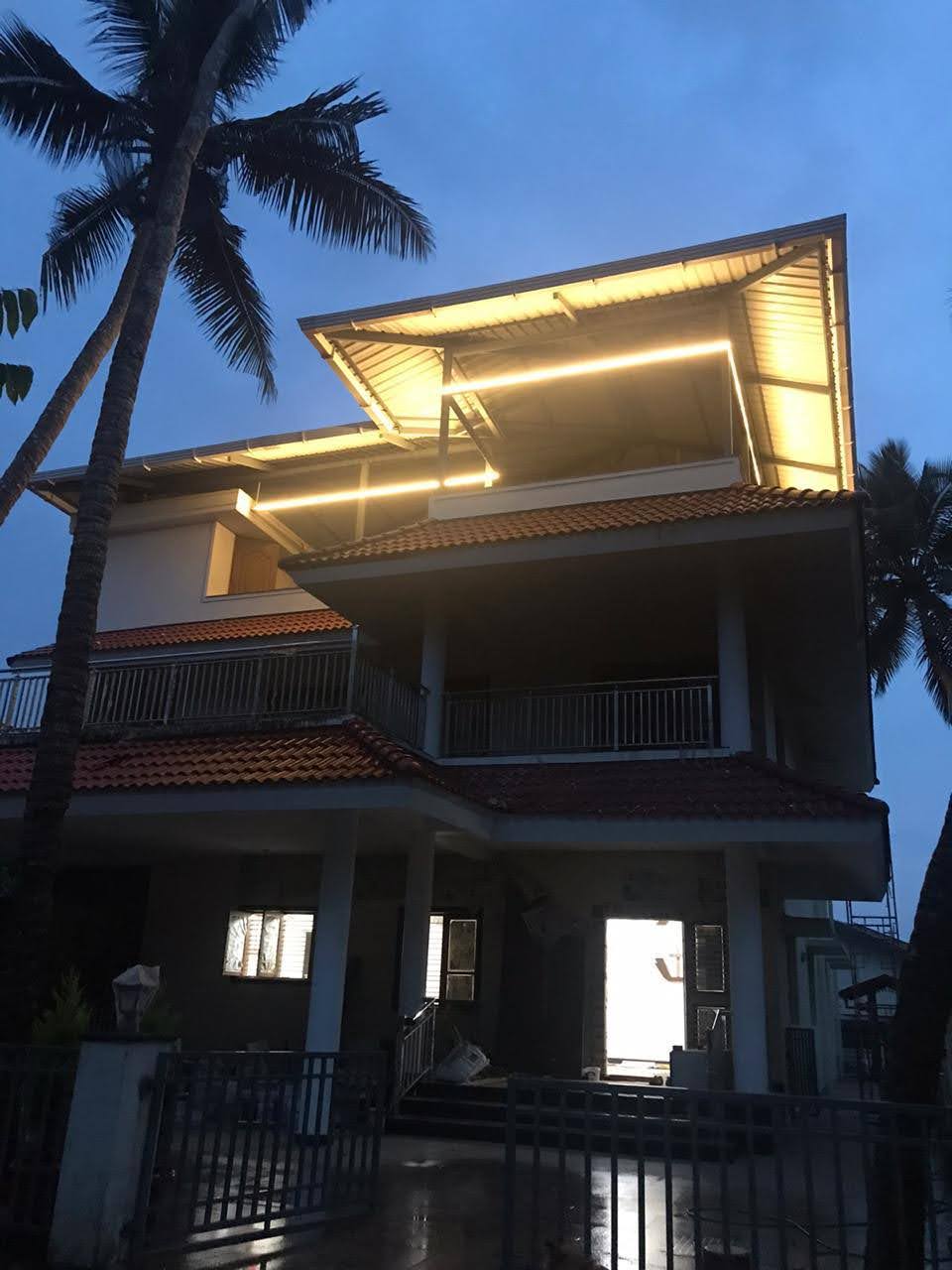 The three-story house features a modern design, highlighted by illuminated roofing. Large windows can be seen, reflecting the evening light, and palm trees frame the structure, enhancing its tropical setting. A gated entrance offers access to the entrance area.