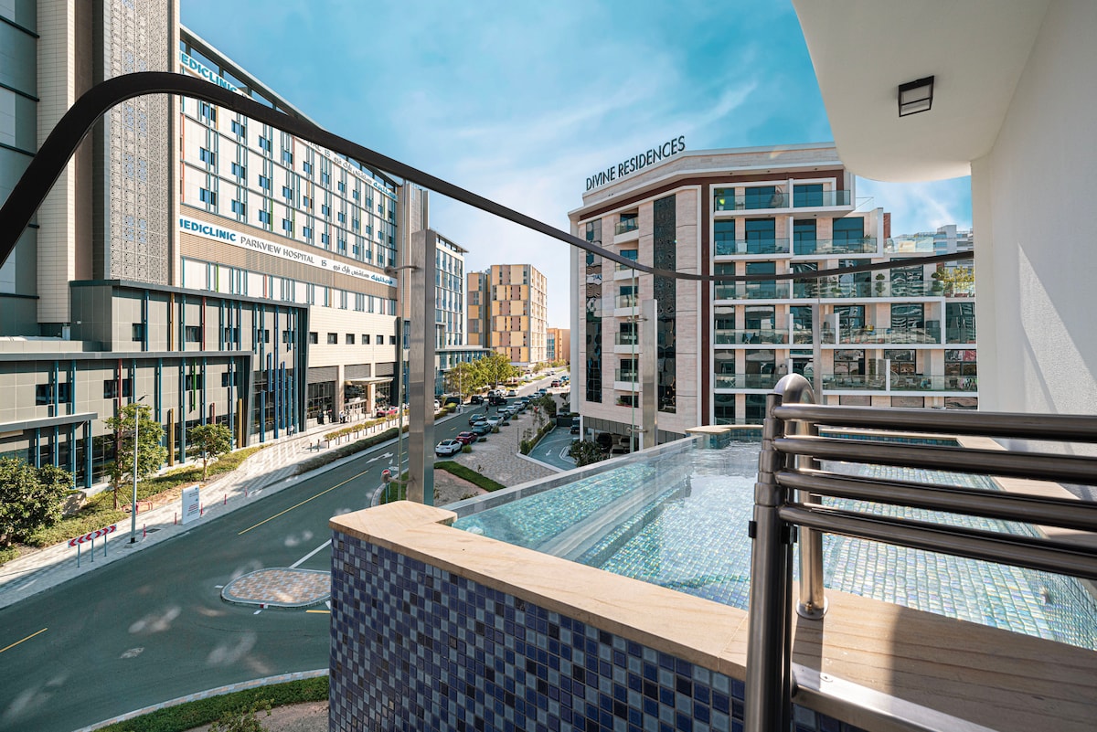 A view from the balcony overlooks a serene private pool, while modern buildings provide a backdrop. The pool features blue mosaic tiles and is framed by a balcony railing. The surrounding area includes sidewalks and greenery, enhancing the urban setting.