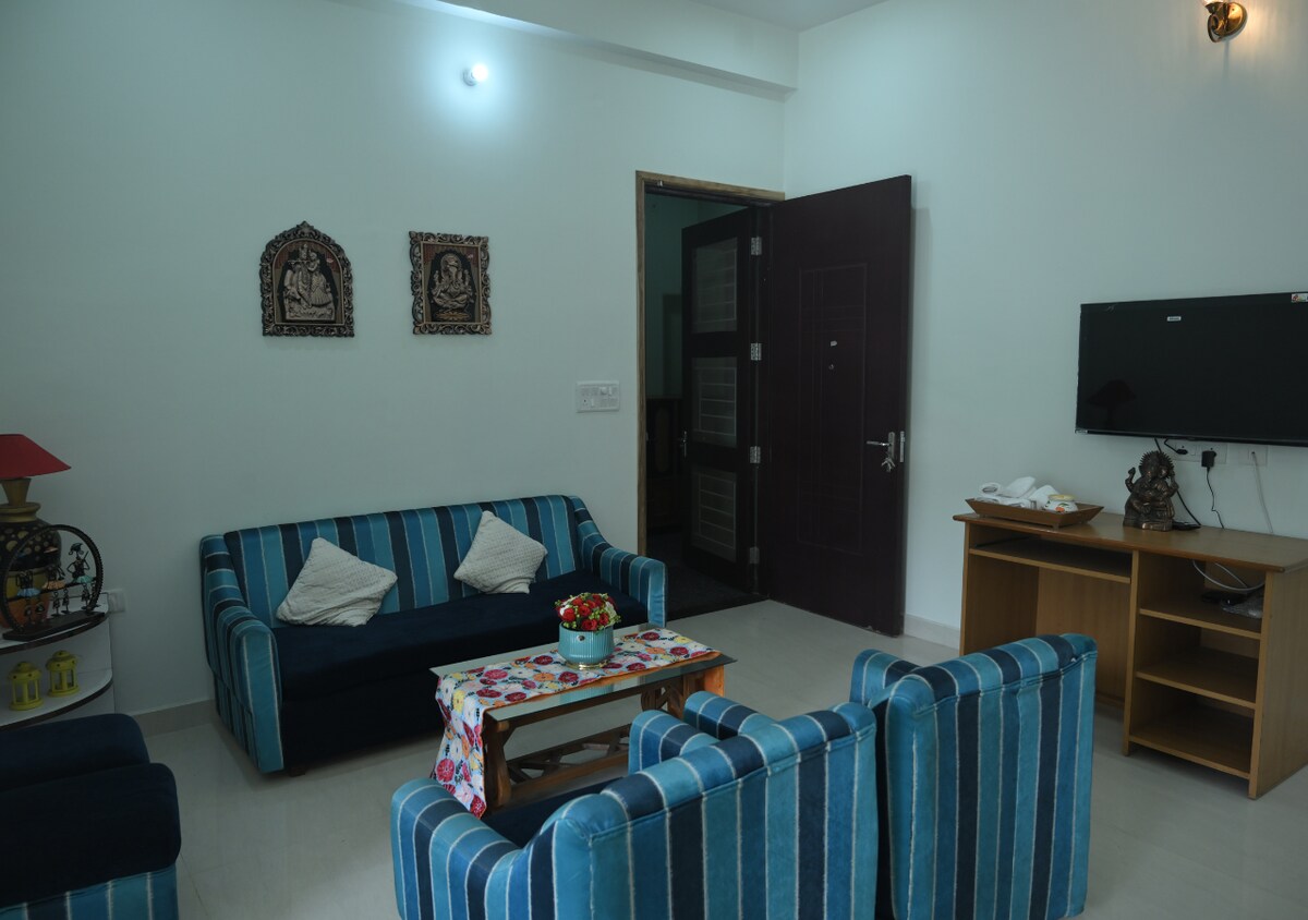 The living area features a combination of blue-striped sofas arranged around a wooden coffee table. Decorative wall art is displayed above, while a television is mounted for entertainment. A sideboard holds additional items, and natural light enters through the doorway.