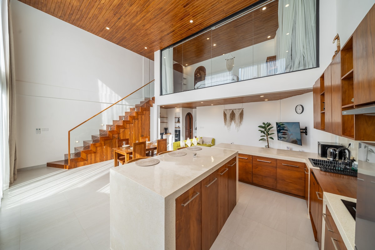 An open-plan kitchen and living area feature modern cabinetry and a large central island. Natural light floods the space through expansive windows, while the dining area is visible in the background. A wooden staircase leads to a lofted area, enhancing the home's spaciousness.