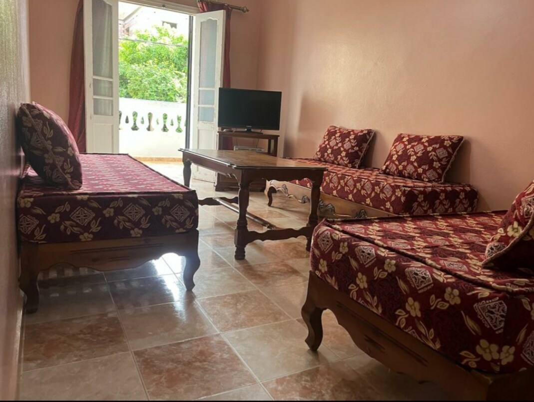 A living area is furnished with patterned seating and a low wooden table. A television is positioned against the wall, and natural light fills the space through open double doors that lead to a balcony, revealing greenery outside.