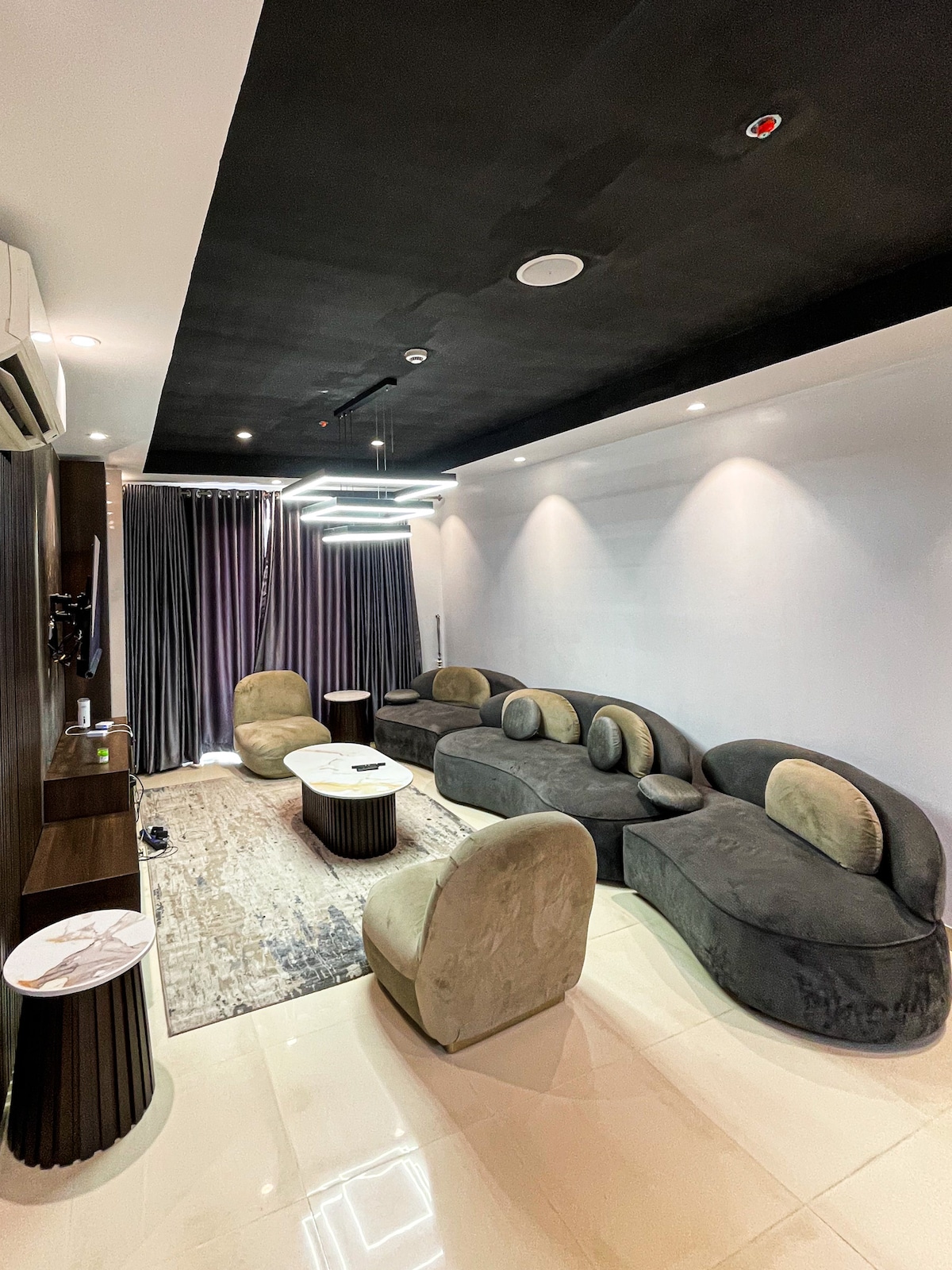 A stylish lounge area is furnished with modern, curved couches in neutral tones. Two small round tables are positioned between the seating. Soft, ambient lighting is provided by a unique overhead fixture, and floor-to-ceiling curtains frame a window along one wall.