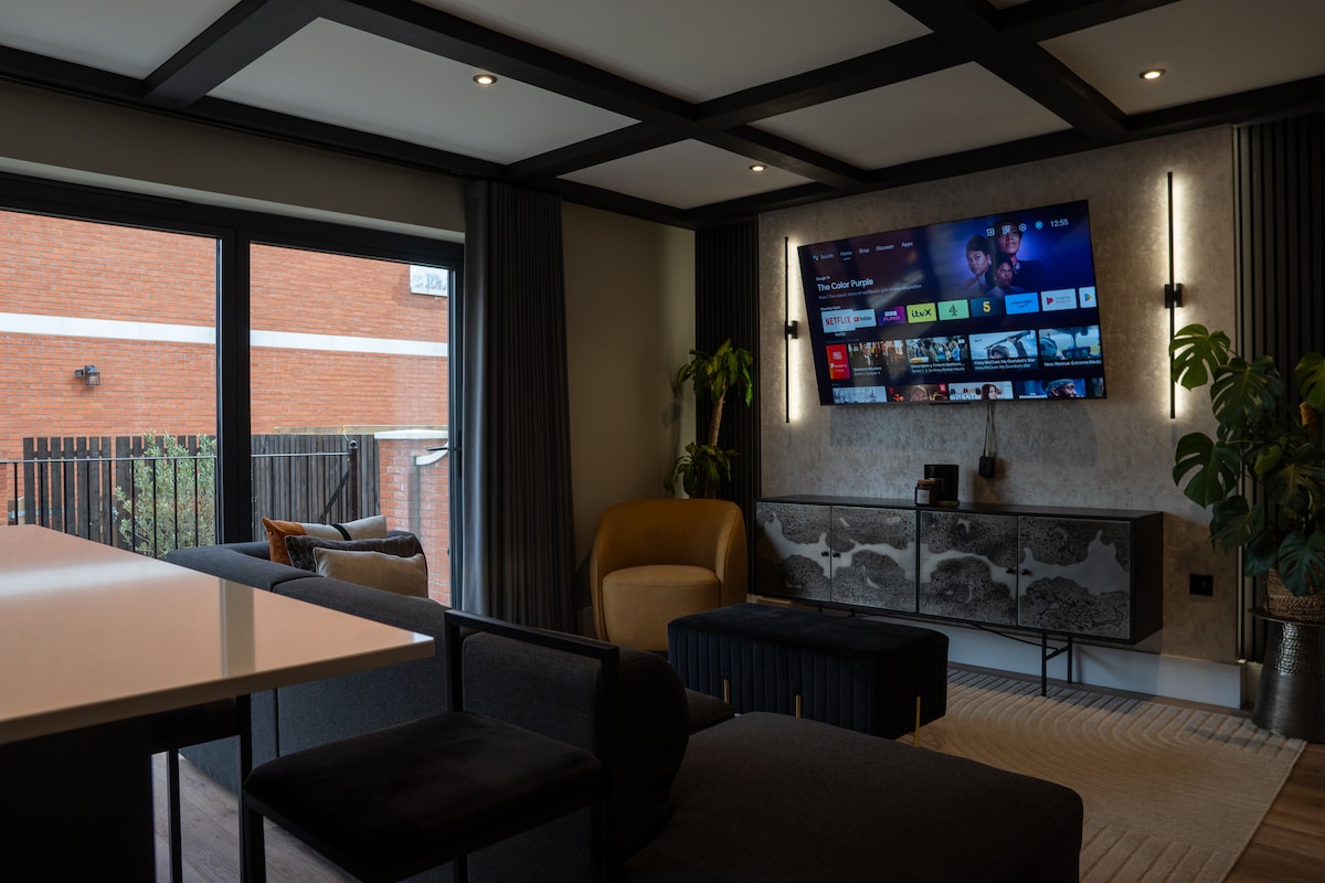 The living area features a large wall-mounted Smart TV, surrounded by dark curtains and modern furniture. Ample natural light enters through sliding glass doors, offering views of the outdoor space. A mix of seating options includes an armchair and sleek couches, inviting relaxation.