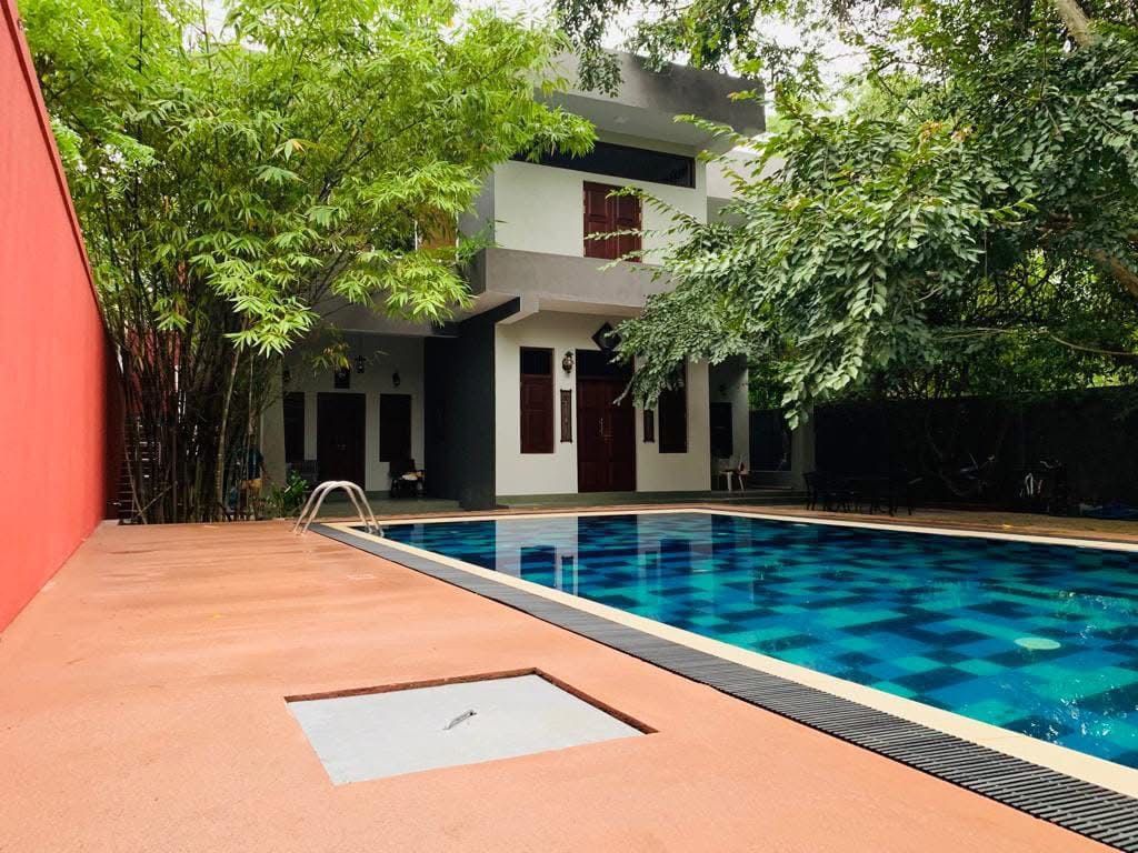 A spacious outdoor area features a sparkling blue pool surrounded by green foliage. The modern villa, with its sleek design, is visible in the background. Comfortable seating is provided near the pool, creating a relaxing atmosphere for guests.