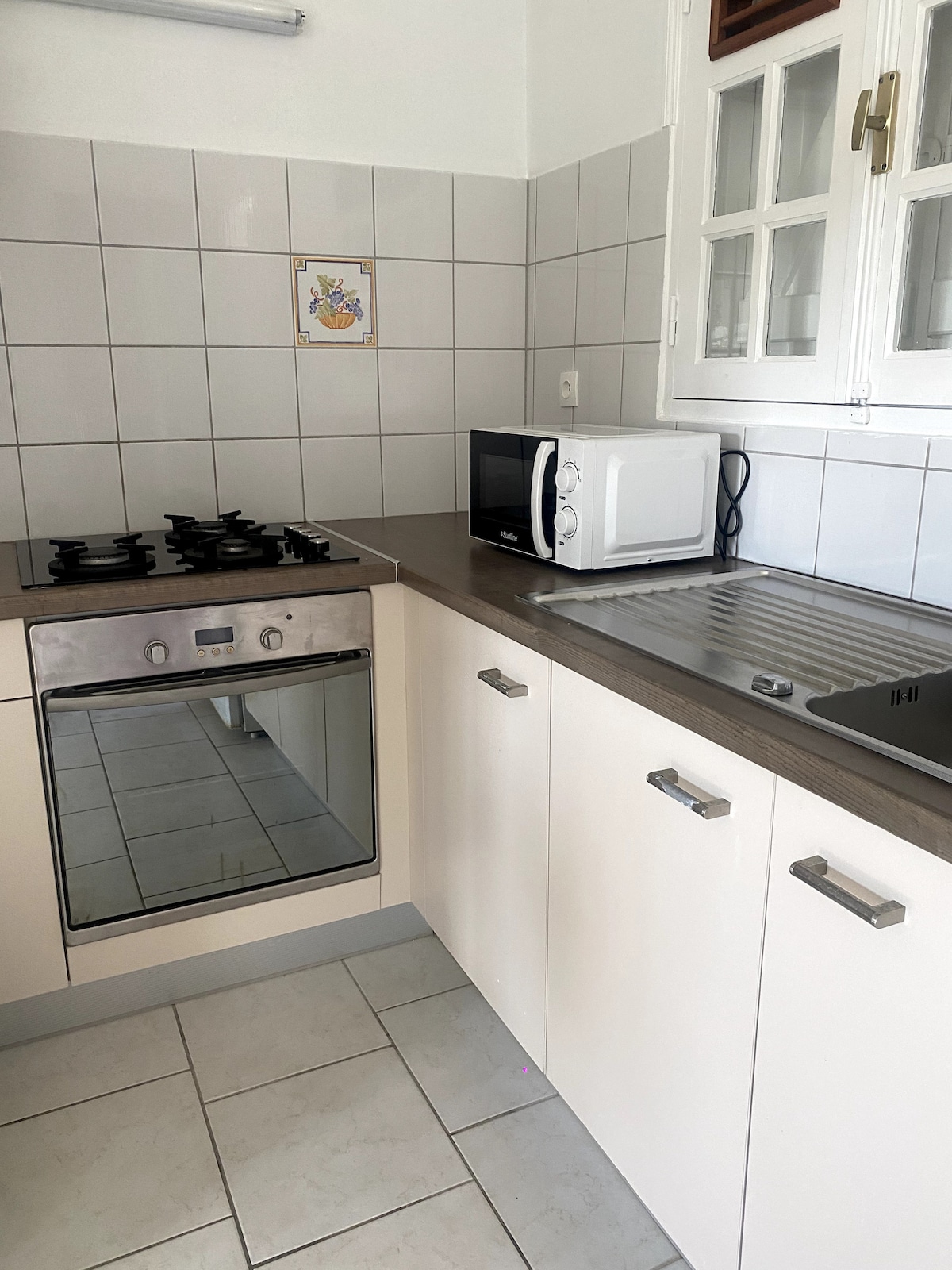 A functional kitchen is displayed, featuring a gas stove, an oven, and a microwave on a countertop. Neutral-colored cabinets and tiled walls create a clean environment. A stainless-steel sink is positioned adjacent to the cooking appliances, with ample countertop space for meal preparation.