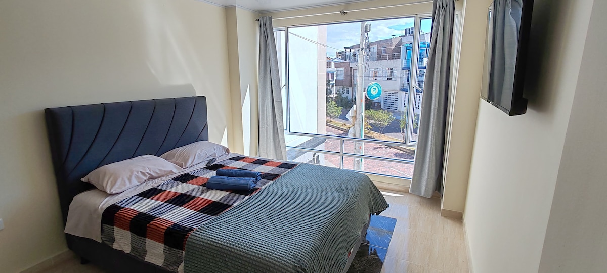 A bright sleeping area features a spacious bed dressed in a patterned blanket, complemented by a soft towel. Large windows allow natural light to fill the room and provide views of the surrounding neighborhood, while curtains add privacy when needed.