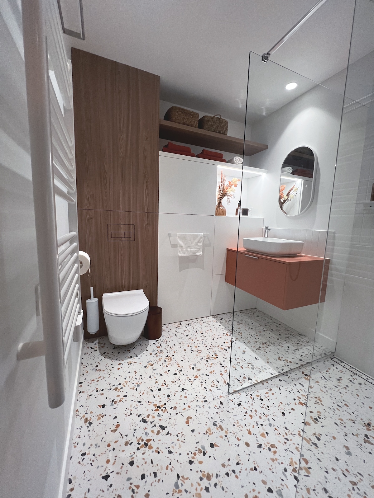 A modern bathroom features a walk-in shower with a glass partition. A round mirror hangs above a sleek vanity with an orange accent, complementing the light wooden cabinetry. The floor showcases a vibrant terrazzo pattern, and a towel is neatly displayed on a rack.