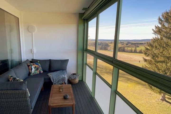 A bright winter garden features large glass windows offering expansive views of the surrounding greenery. A cozy seating area is set up with a sofa and decorative cushions, accompanied by a small wooden coffee table. The serene landscape outside is visible, enhancing the tranquil atmosphere.