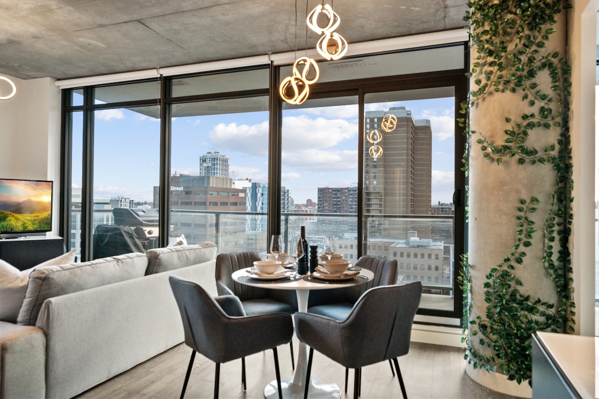 A modern dining area features a round table set for two, accompanied by four stylish chairs. Floor-to-ceiling windows provide expansive views of the downtown skyline, while decorative lighting and greenery enhance the ambiance of the space.
