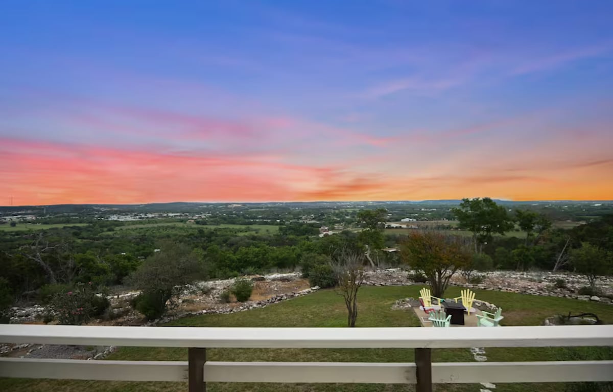 A spacious outdoor area reveals a scenic view of the Hill Country at sunset, with vibrant colors painted across the sky. A gathering space with seating is visible, set against a backdrop of rolling hills and open greenery.