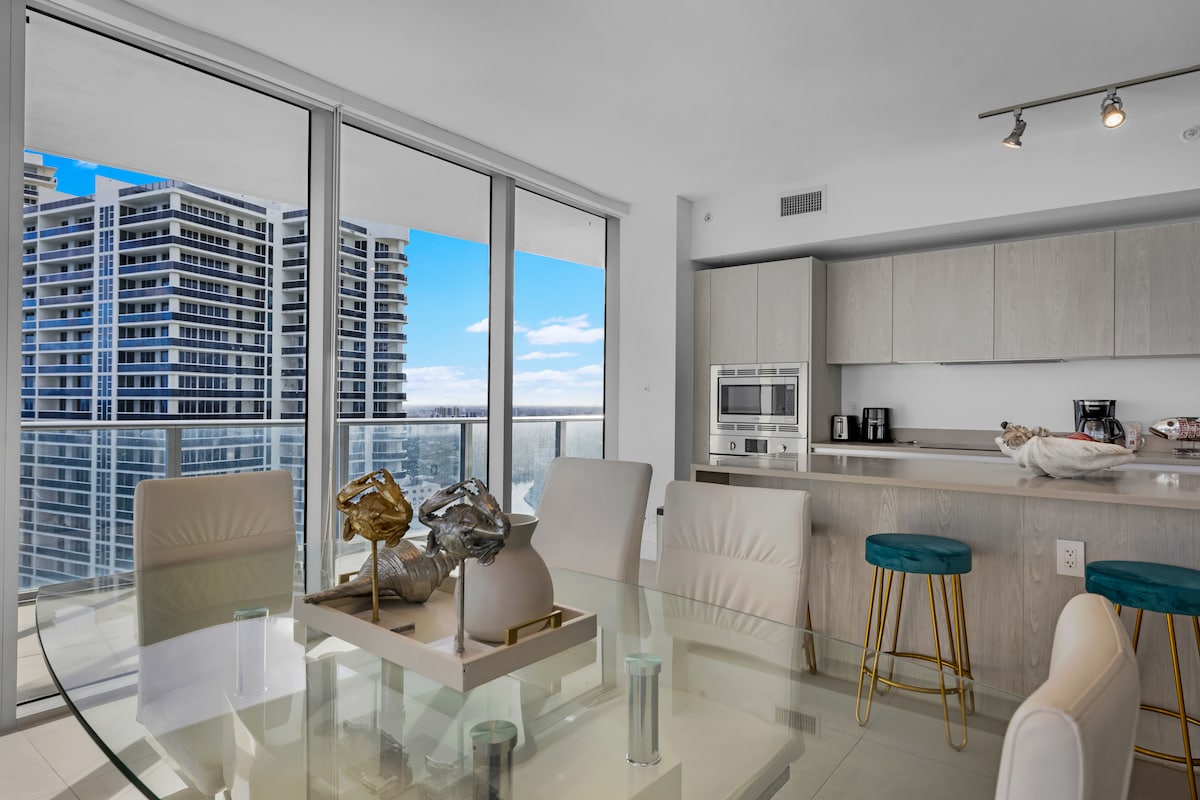 A bright dining area features a glass table surrounded by white chairs, complemented by teal bar stools. Large windows provide views of the city skyline, allowing natural light to fill the space. A minimalist kitchen area is visible, showcasing modern appliances.