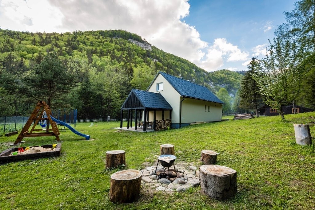 An outdoor area features a stone fire pit surrounded by log seating. A spacious cabin sits in the background, framed by trees and mountains. A playground with a slide is visible, alongside a sand area, creating an inviting space for children to play.