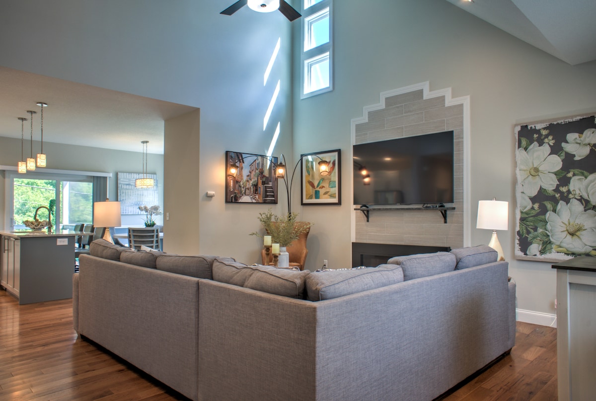 A spacious living room is highlighted by a large sectional sofa facing a wall-mounted television. Natural light filters through tall windows, casting soft shadows. Decorative artwork adorns the walls, and a modern fireplace is positioned below the television.