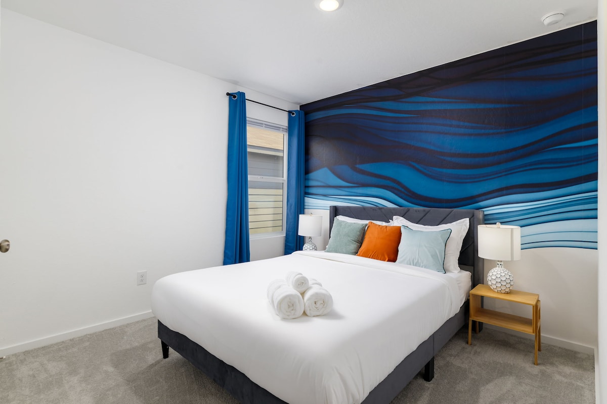 A bedroom features a spacious bed with crisp white linens and decorative pillows in shades of blue and orange. A captivating hand-painted mural in blue waves adorns one wall, while bedside tables with lamps provide soft lighting. Light filters through a window with blackout curtains.