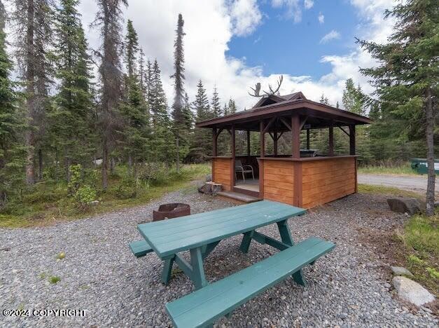 An outdoor gazebo features a wooden structure with a sloped roof, surrounded by trees. A picnic table painted in turquoise is positioned nearby on a gravel surface. Natural scenery frames the area, providing a serene setting for gatherings.