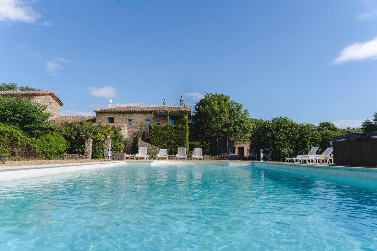 A large swimming pool is visible, featuring clear turquoise water and surrounded by greenery. Several white lounge chairs are positioned around the pool's perimeter, with a rustic stone building in the background and a clear blue sky overhead.