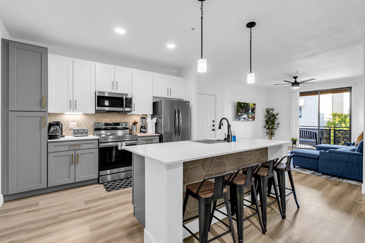 A fully equipped kitchen is showcased, featuring modern appliances and sleek cabinetry. A spacious island with barstools is complemented by warm, ambient lighting. Natural light flows in from a nearby window, illuminating the open layout that connects to a cozy living area.