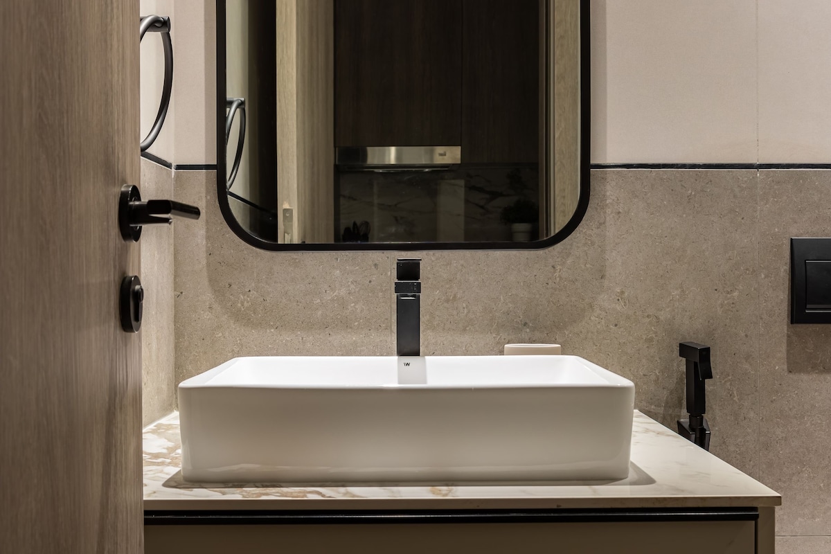 A modern bathroom is presented, featuring a sleek, rectangular sink atop a minimalist countertop. A round mirror is mounted above, reflecting the space, while dark fixtures complement the light stone walls and give a contemporary feel.
