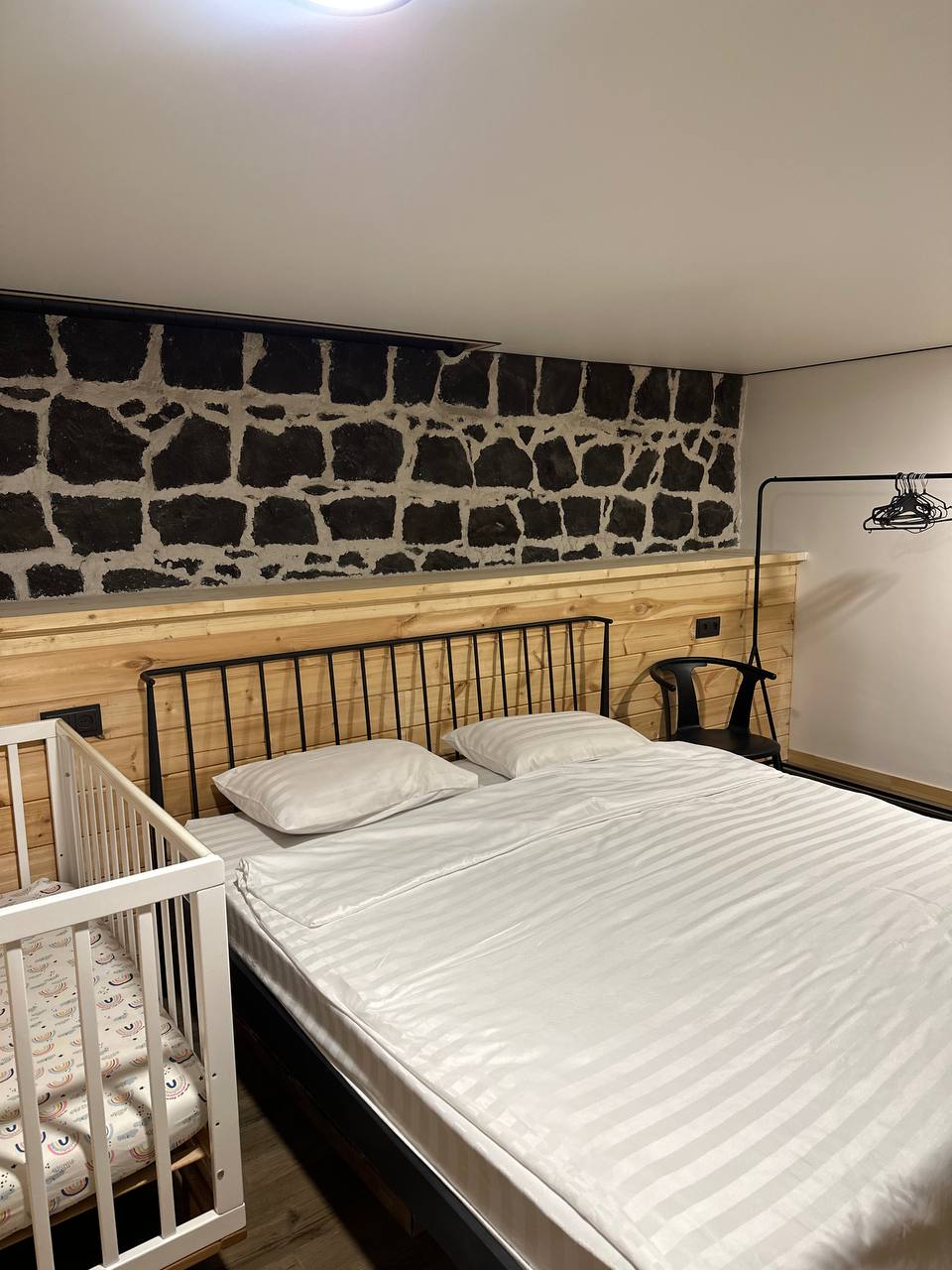 A sleeping area is presented with a spacious bed featuring white linens. Adjacent, a crib is visible, designed in a neutral tone. A stone accent wall adds texture, complemented by wooden elements. A clothing rack stands nearby, contributing to the room's functional layout.