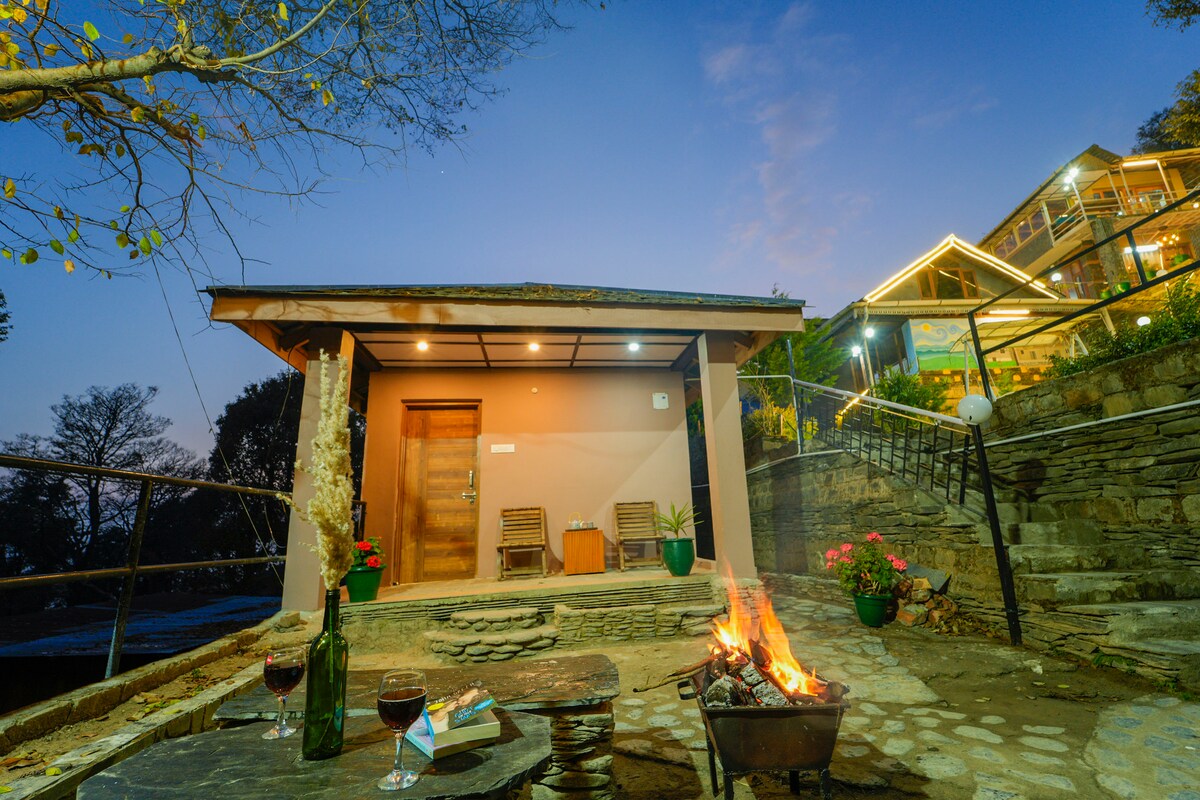 A charming outdoor space is showcased, featuring a fire pit surrounded by seating. The entrance is marked by wooden doors, leading to a cozy building. Soft evening light casts a gentle glow, highlighting stone steps and planted greenery, contributing to a serene ambiance.